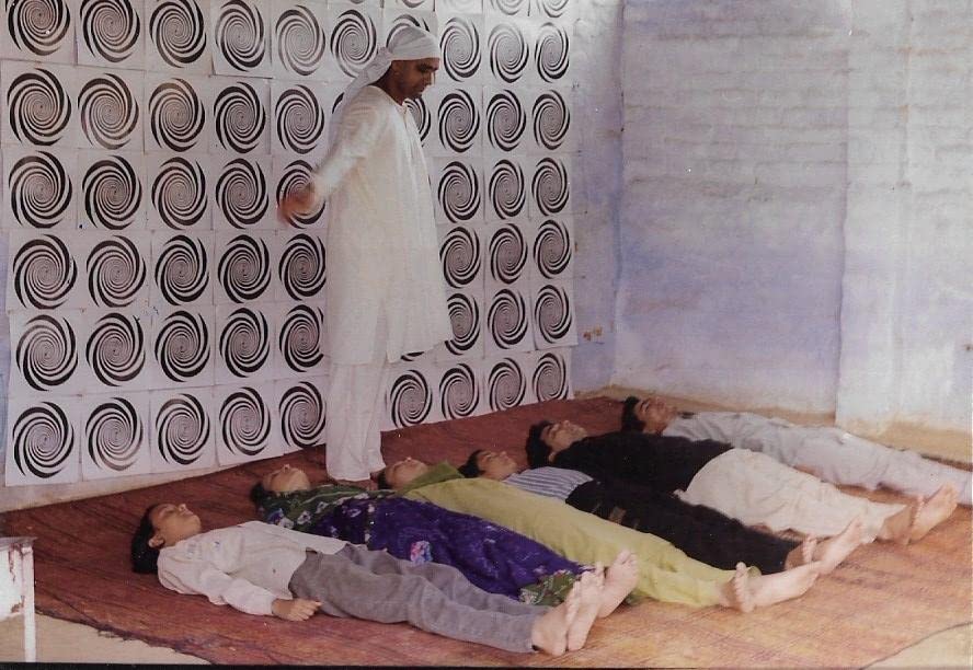 A practitioner in white attire conducting a group hypnosis or yoga nidra session with participants lying on mats in a serene room with spiral-patterned walls.